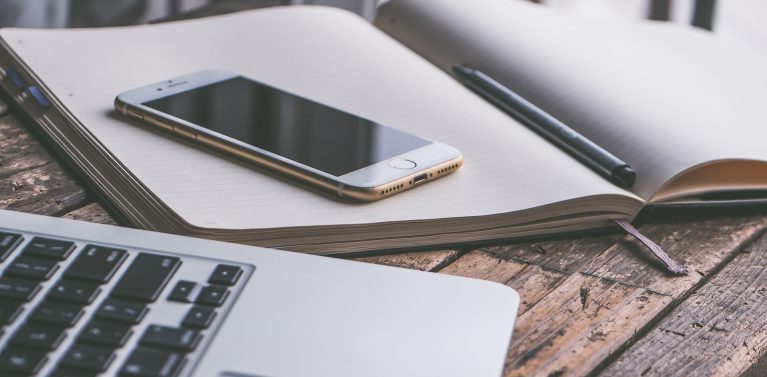 image of a notepad open with a gold cased iphone and a pen used as a paper weight on a rustic wooden table.