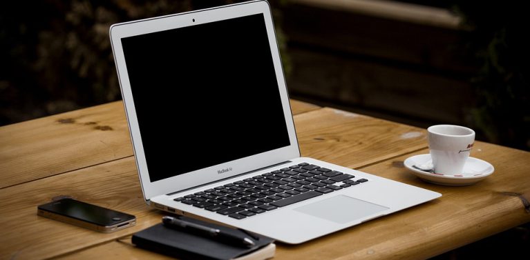 image of a laptop on an oak table with a coffee to the right and a phone with a notepad and pen on the left.