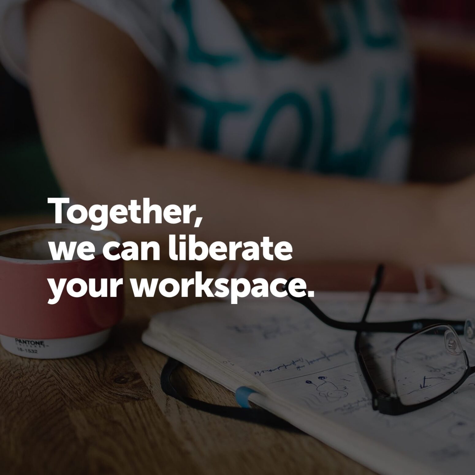 This image features bold white text stating "Together, we can liberate your workspace." overlaid on a blurred background showing a workspace scene. The background appears to be a wooden desk with a notebook containing handwritten notes, a pair of eyeglasses, and a cup or mug